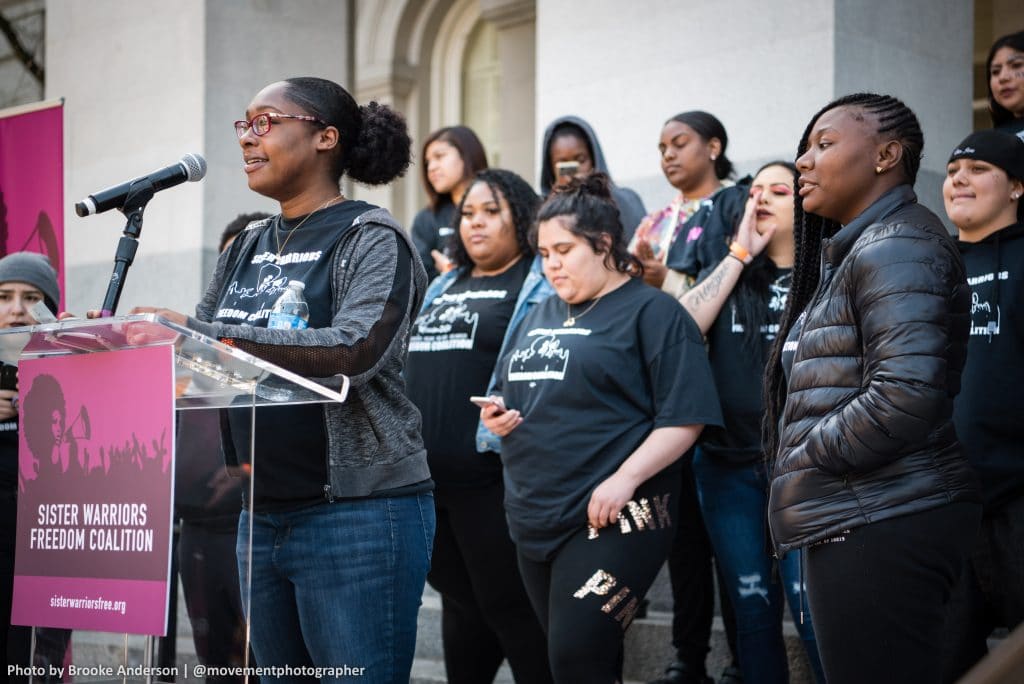 A young Black woman with glasses wearing a jacket and jeans speaks at a podium with 9 young women standing behind her