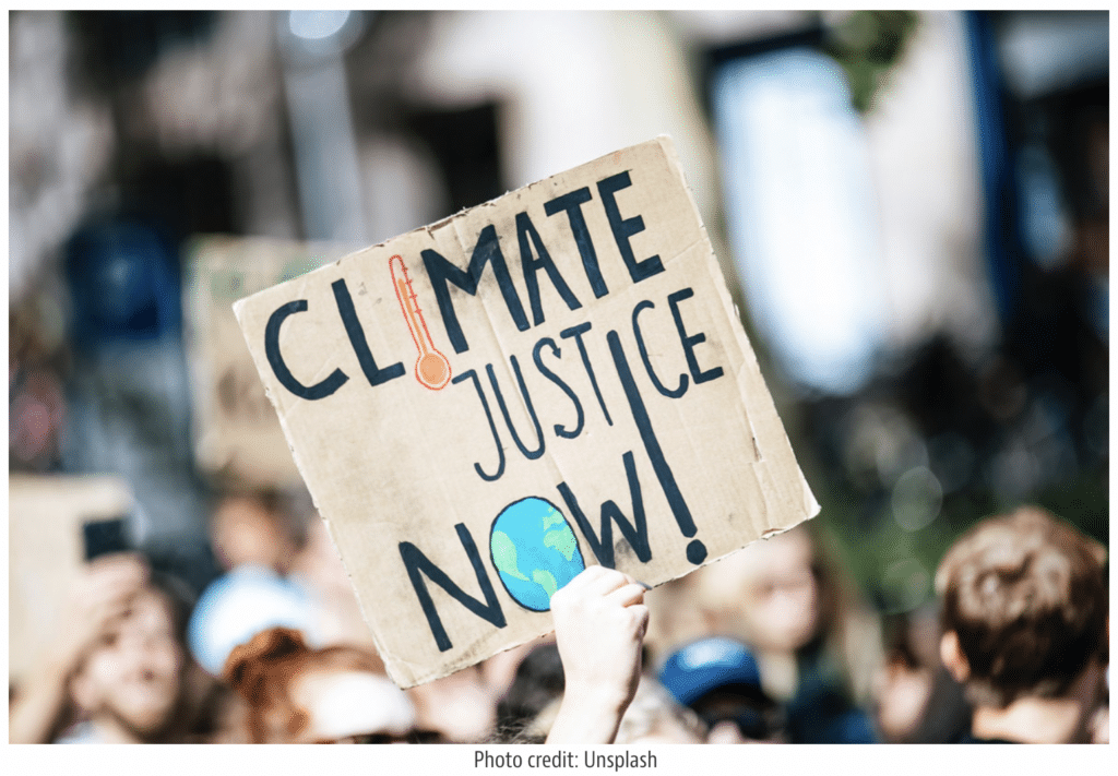 Hand holds up protest sign that reads "Climate Justice Now!" in a crowd.