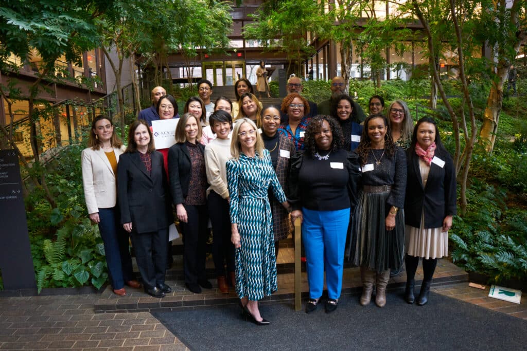 The New York Small Business Funders Collective and the five inaugural grantee partners stand together for a group photo with plants in the background. 