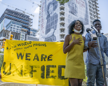 A person with brown skin and black hair wears a yellow dress and speaks into a microphone in front of a yellow sign that reads 