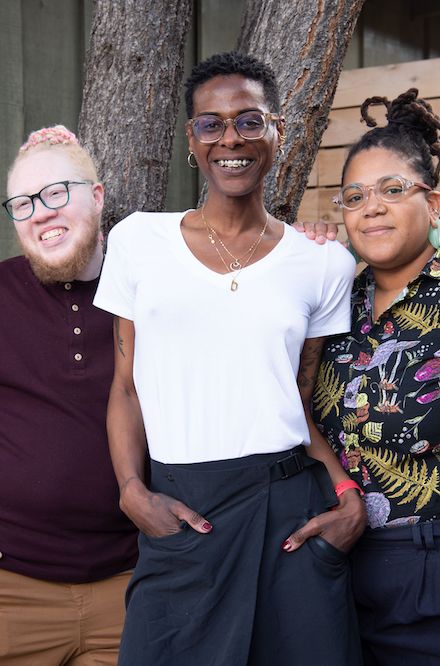 Person with light skin and ginger hair wears glasses and a burgundy shirt. Bre Rivera center with brown skin and short black hair. She is wearing a white shirt. Person on the right with light brown skin and black hair worn in dreadlocks up in a bun.