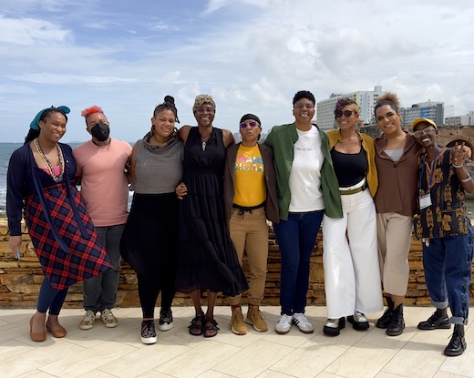 Nine people stand arm-in-arm on a board walk in front of the ocean and smile