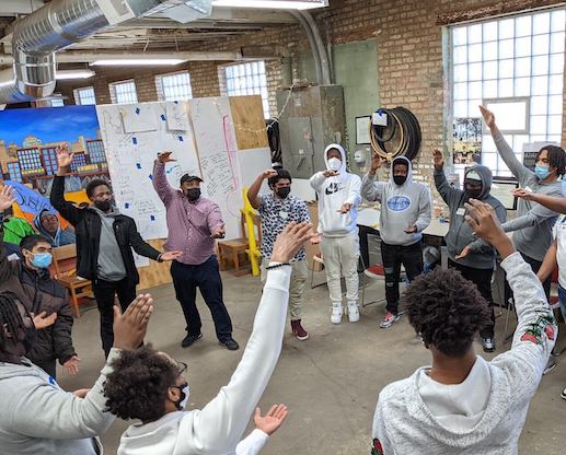 Students stand in a circle doing an exercise with one arm up and one arm in front of them.