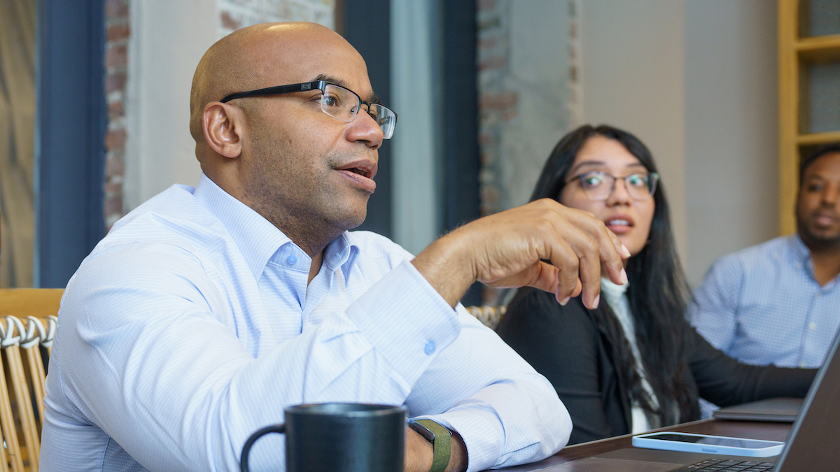 Person with brown skin and a shaved head wears glasses and a blue button-down shirt. He sits at a table with a couple other people in front of computers and talks.