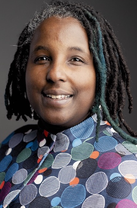 Lauren Jacobs headshot - person with brown skin and black chin-length dreadlocked hair wearing a purple and blue polka dotted blouse.