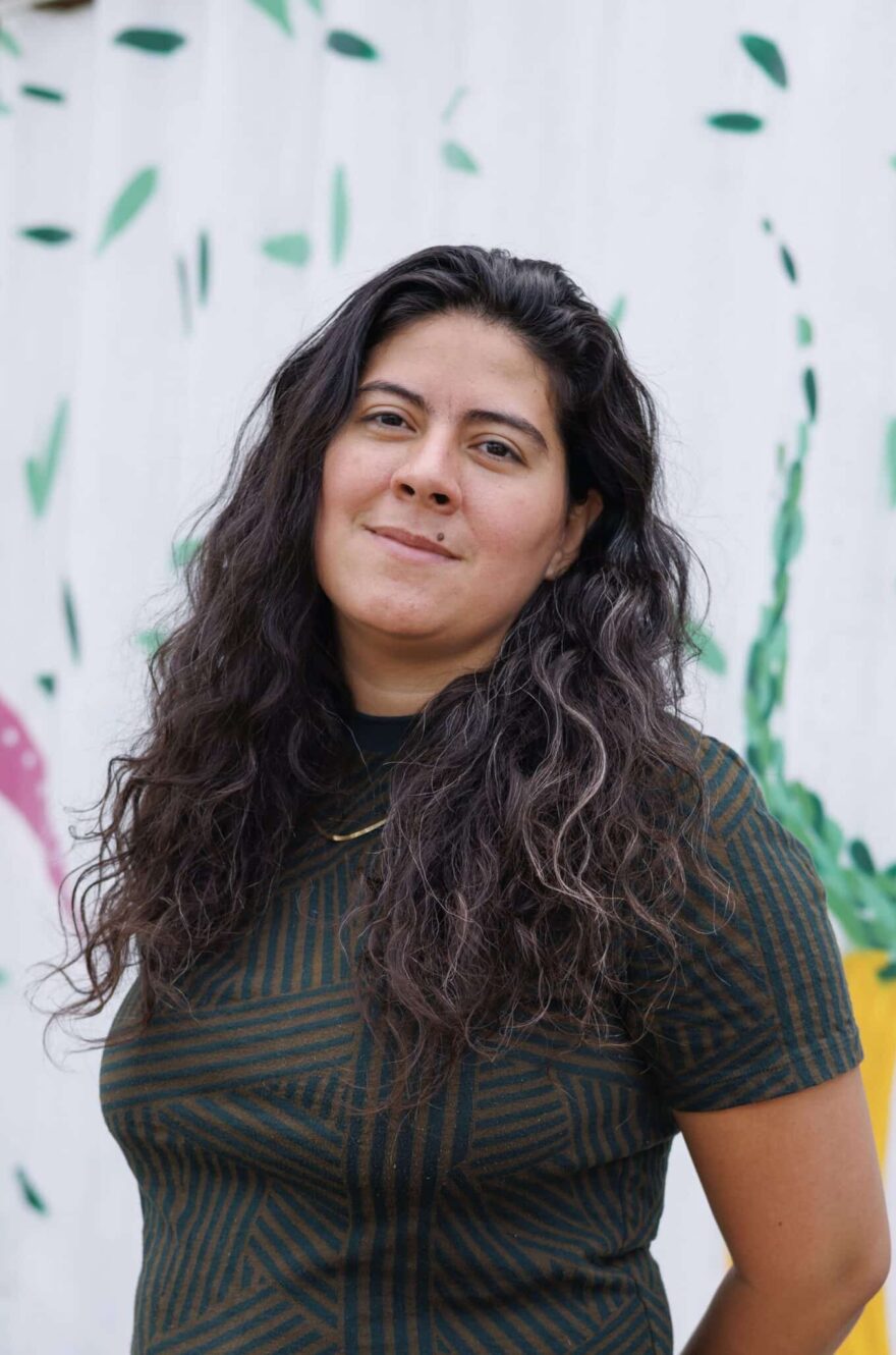 Maria Lopez- Nuñez headshot - person with tan skin and long black wavy hair wears a black t-shirt standing in front of a colorful mural.