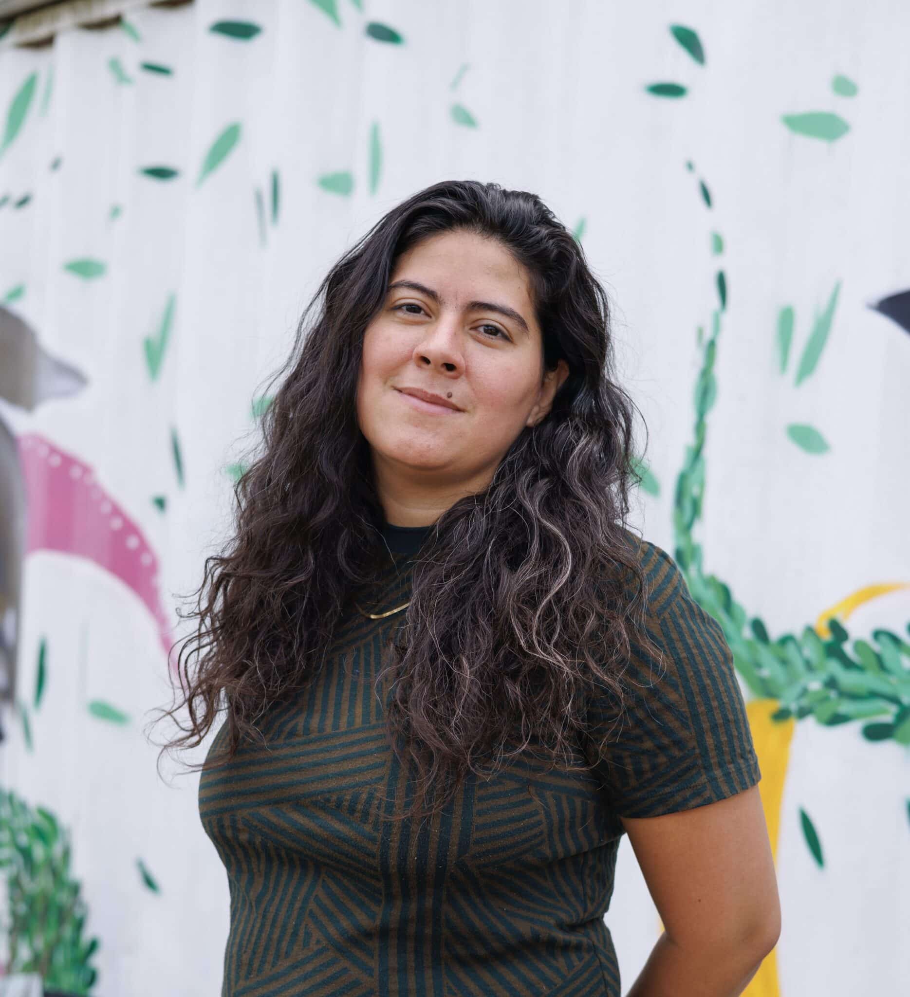 Maria Lopez- Nuñez headshot - person with tan skin and long black wavy hair wears a black t-shirt standing in front of a colorful mural.