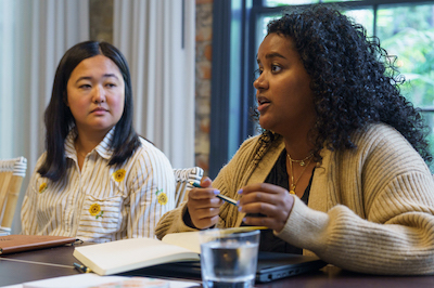 One person with brown skin and curly black hair sits at a table and speaks with a person with light skin and shoulder-length black hair.