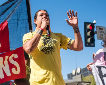 Indigenous person wearing a yellow shirt speaks into a microphone to a crowd in the street