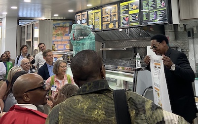 A group of people stand together in a bodega and a man with a poster speaks to the crowd