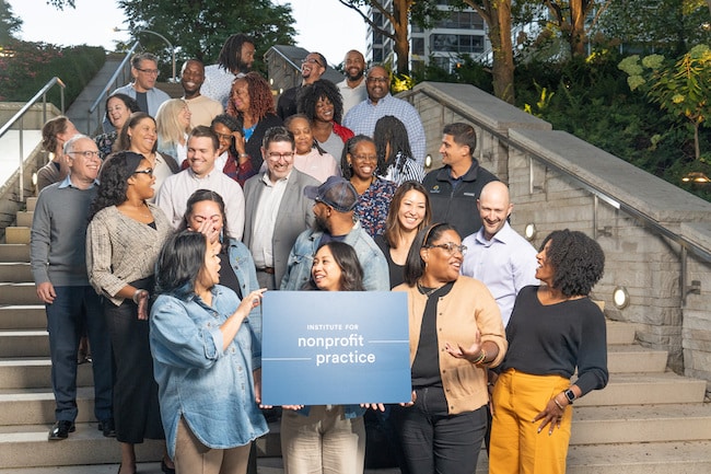 A group of people smile and laugh while standing on steps together holding a sign that reads 