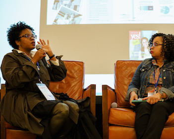 One person with brown skin and short hair sits in a chair on a stage and speak into a microphone. A person with brown skin and should-length curly hair sits next to her and listens.