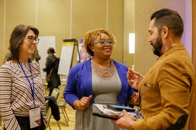 Three people stand speaking to one another. The person in the middle is speaking and has brown skin and short natural hair wearing blue glasses.