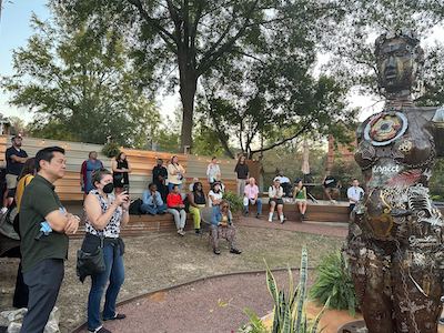 A group of people sit on a low wall in a back garden area and look at a metal sculpture of a Black woman.