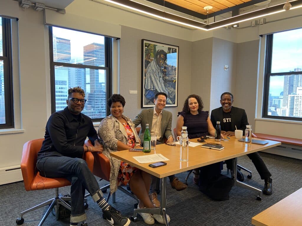 A group of five people sit side-by-side at a table in a conference room and smile for a photo. 