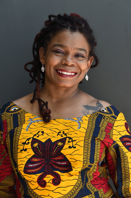 A woman with brown skin and her hair in dreadlocks smiles at the camera wearing a yellow and red dress.