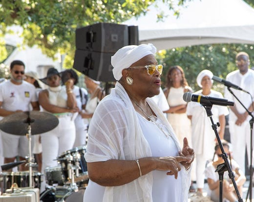 An older woman with brown skin wearing a white turban headscarf, a white dress, and a white shawl, singing in front of a group of people and a band.