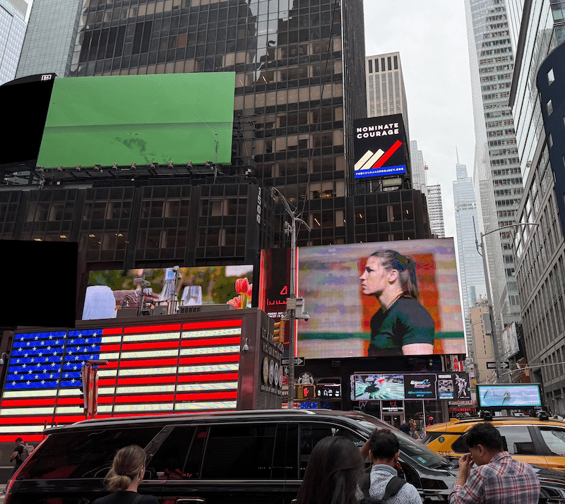 Times Square in New York City - billboards on a busy city street. One billboard reads "Nominate Courage: The Courage Project" 